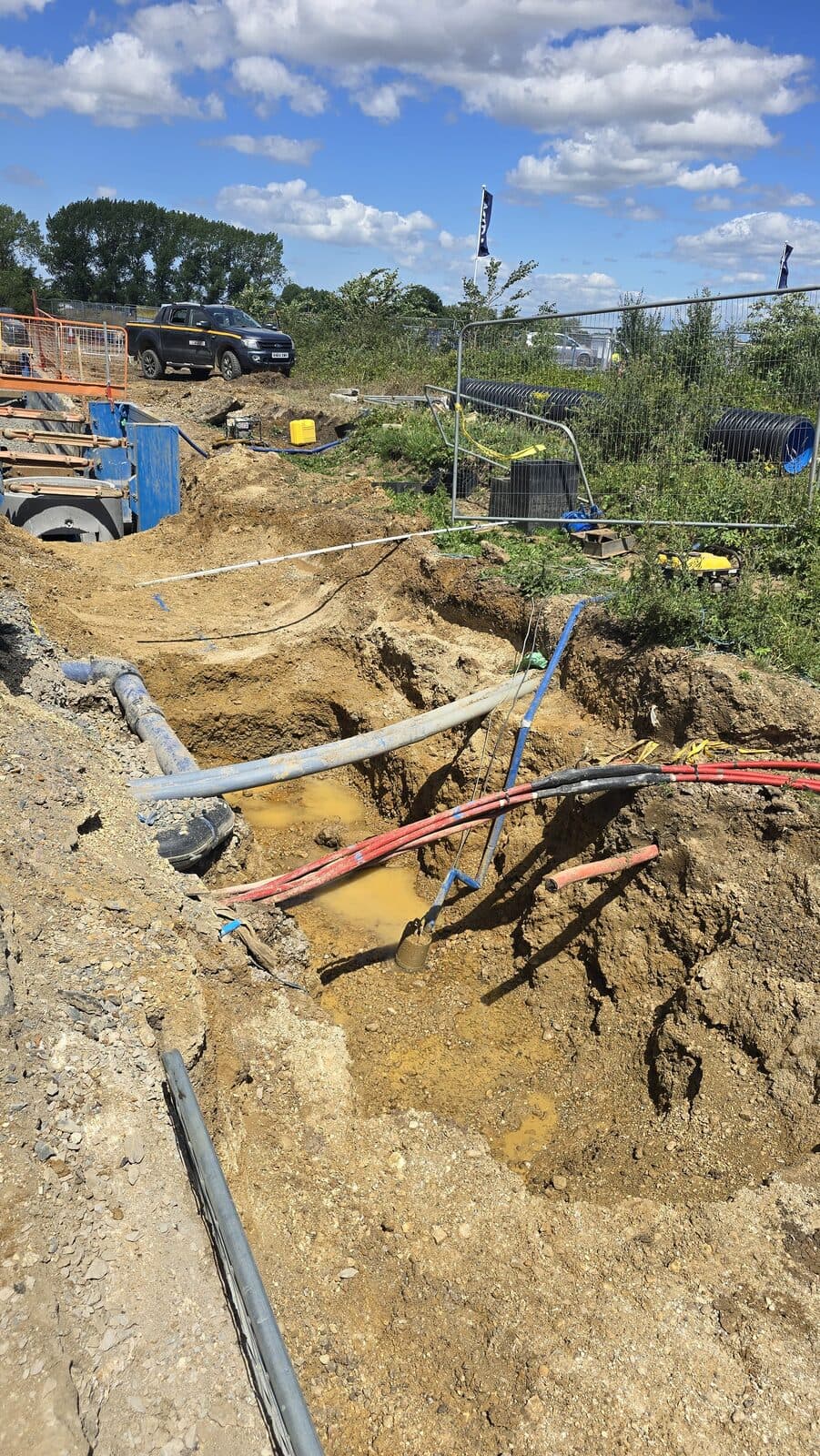 Deep excavation trench exposing multiple utility pipes and red electrical cables with waterlogged base, under blue sky with parked vehicle nearby