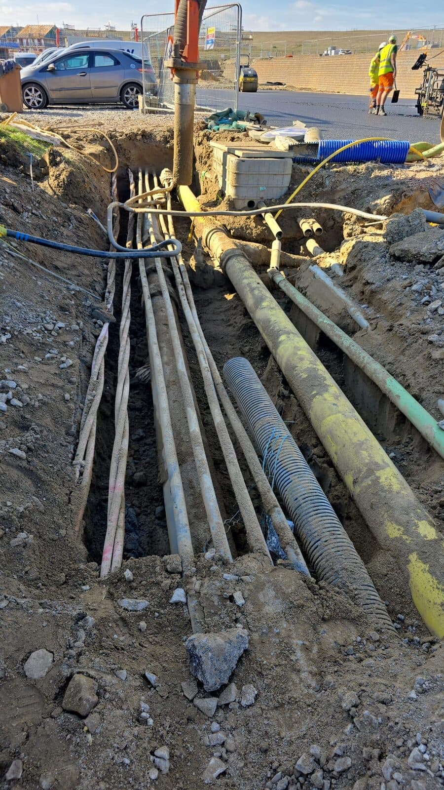 Deep trench showing multiple exposed pipes including yellow ducting and vertical support stakes at residential construction site